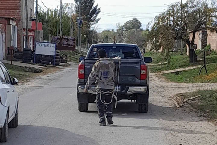 Un hombre de espaldas camina tirando de una soga atada a la parte trasera de una camioneta gris.