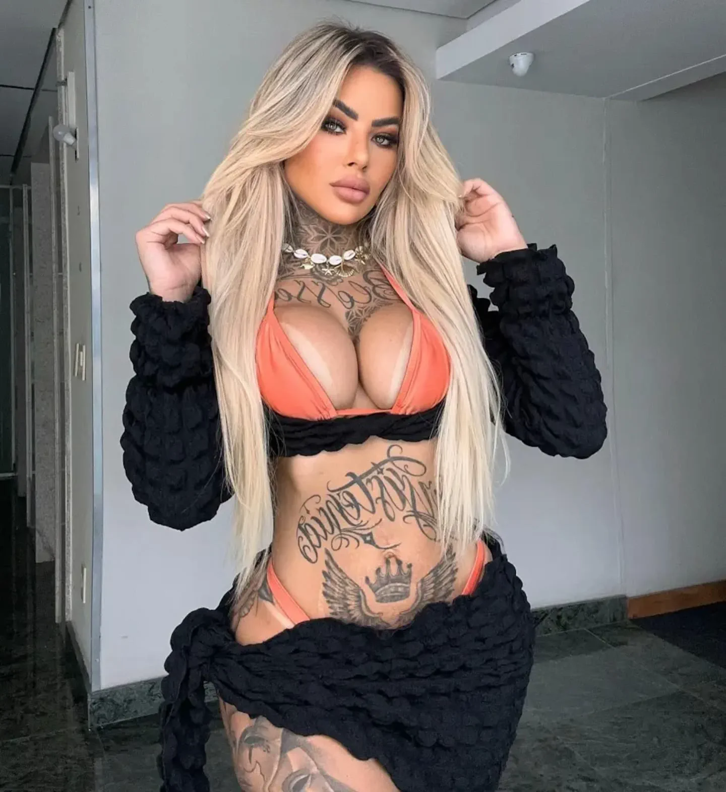 Una mujer rubia y tatuada posa en bikini naranja y un conjunto negro en un interior.