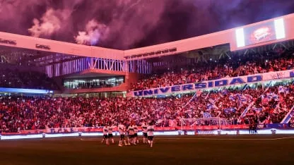Un equipo de fútbol festeja en una cancha de noche frente a una tribuna colmada con banderas y luces violetas.