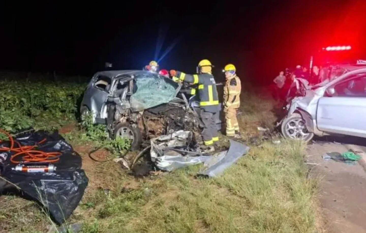 Bomberos asisten a un choque nocturno entre dos autos visiblemente destruidos a un costado del camino.