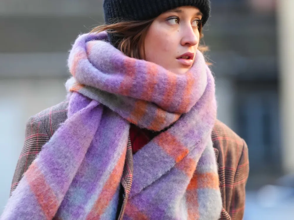 Una joven con gorro de lana negro lleva una bufanda gruesa a cuadros violetas y naranjas al cuello.