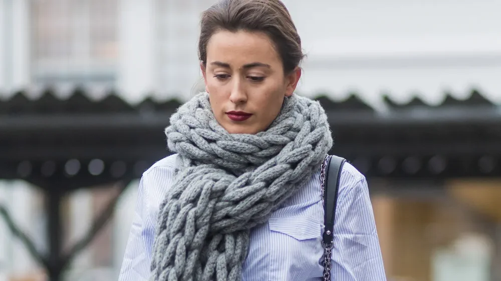 Una mujer con el pelo recogido y camisa a rayas lleva una bufanda tejida gris muy gruesa.