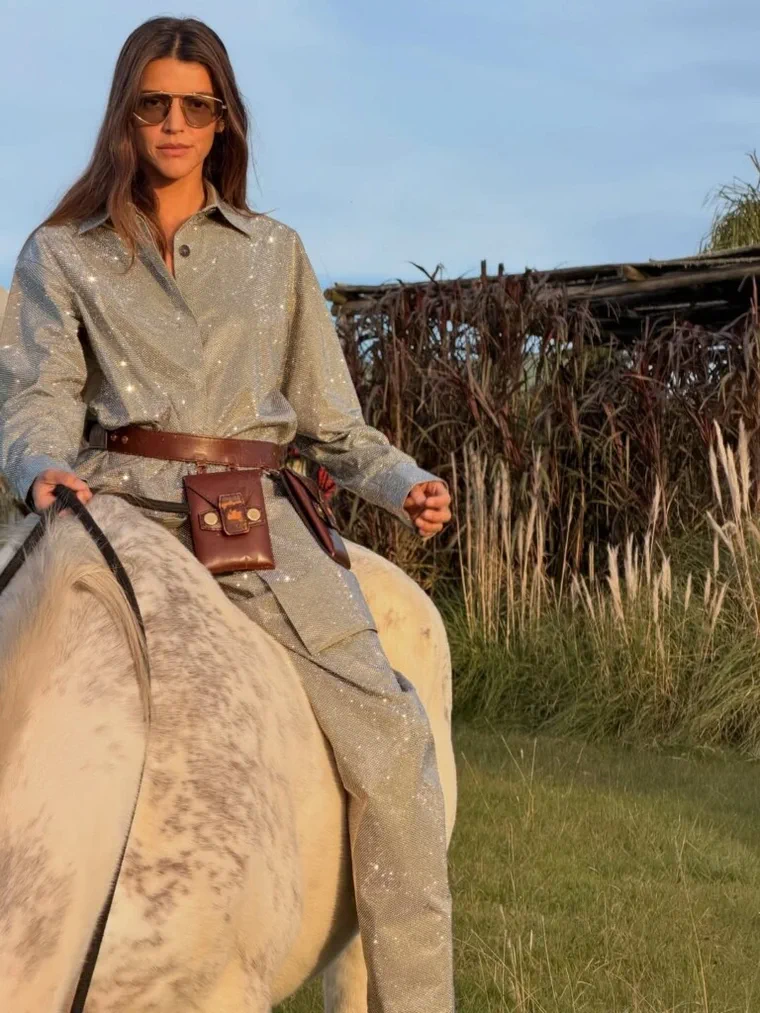 Una mujer con anteojos oscuros y un conjunto brilloso montada a caballo al aire libre.