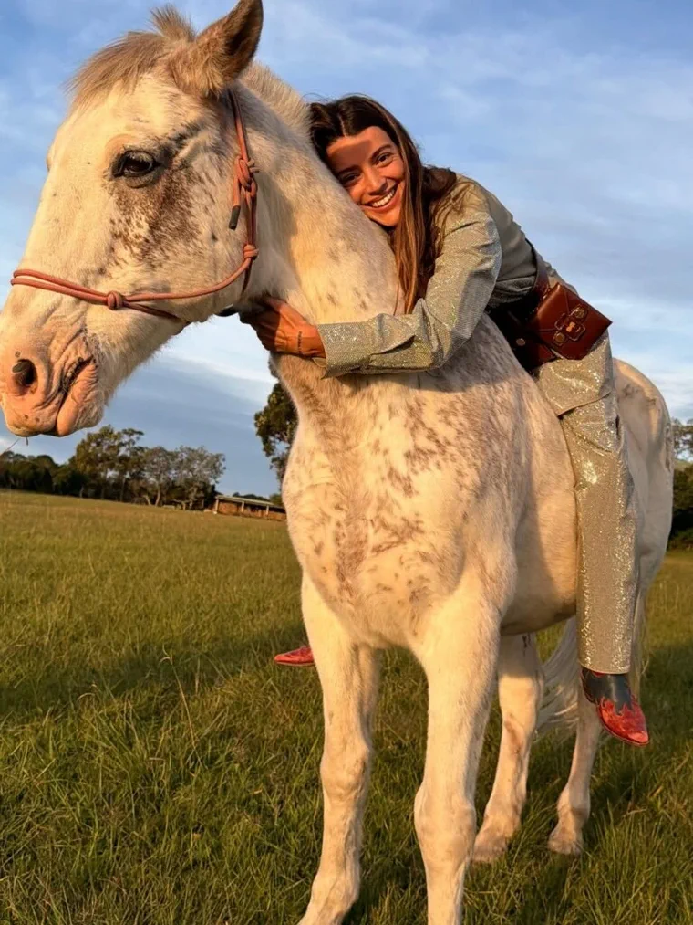 Una mujer sonriente vestida de fiesta abrazando el cuello de un caballo blanco en un pastizal.