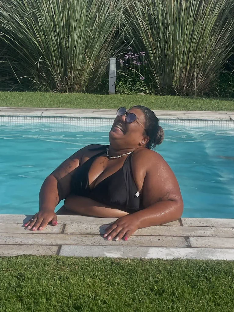 Una mujer con anteojos de sol y bikini negra se apoya sonriendo en el borde de una pileta.