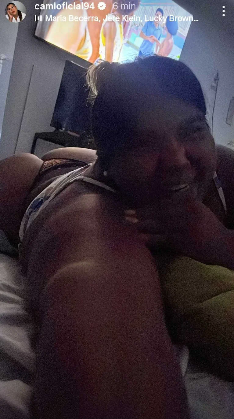 Una mujer acostada boca abajo en una cama sonríe en una habitación mientras se ve un televisor encendido al fondo.