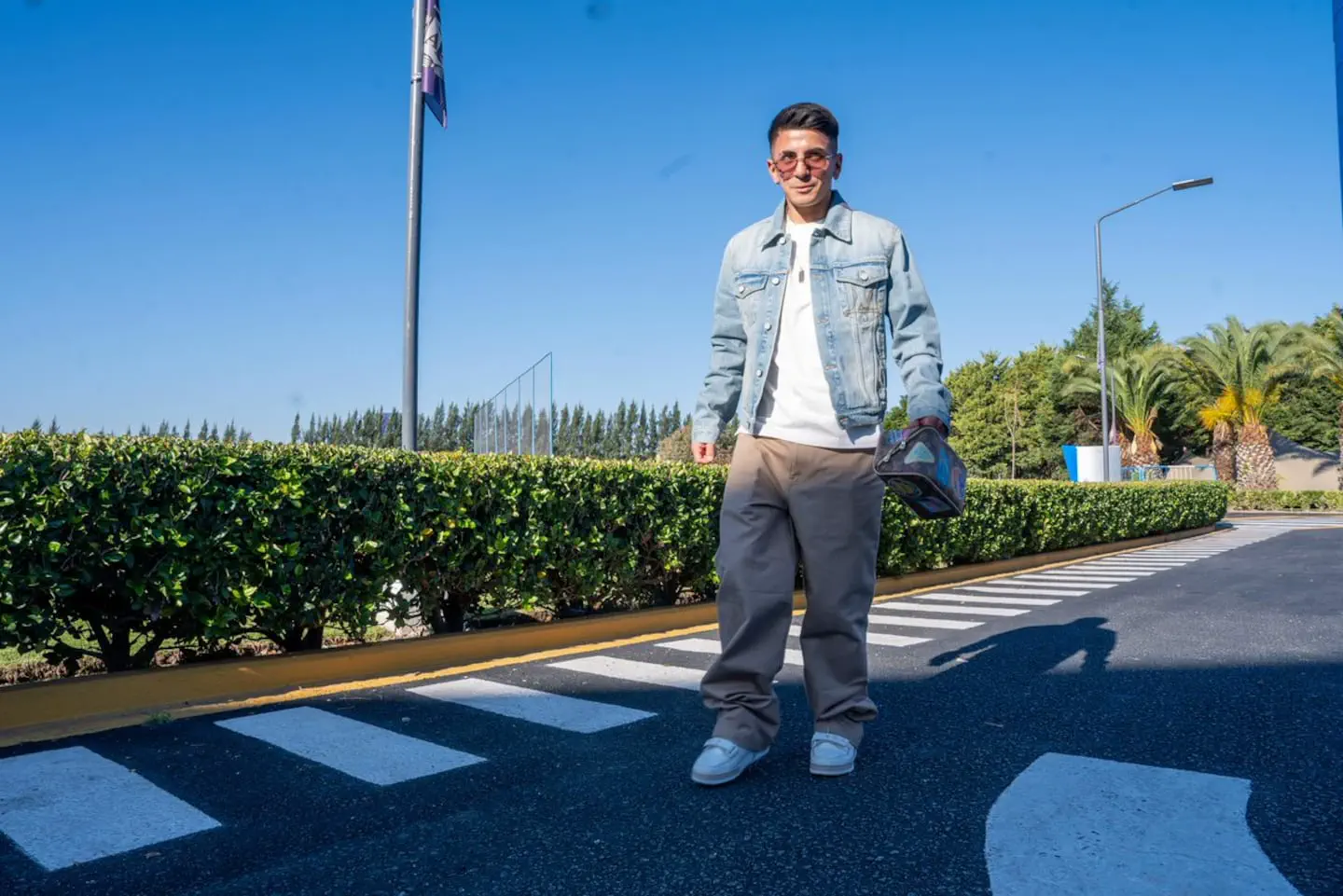 Un pibe con campera de jean, anteojos oscuros y un bolso camina al lado de una calle interna.