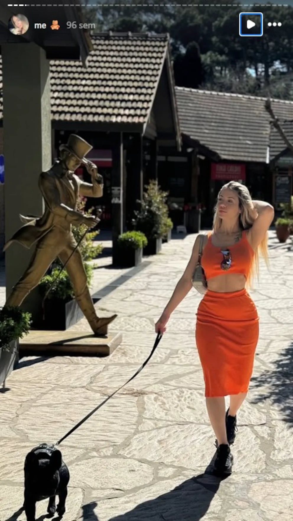 Una chica de conjunto naranja camina con su perro bulldog francés con correa al lado de una estatua.