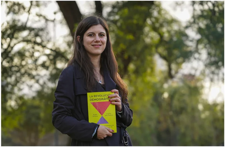 Carola della Paolera posa al aire libre sonriendo y sosteniendo el libro La revolucion demografica con ambas manos.