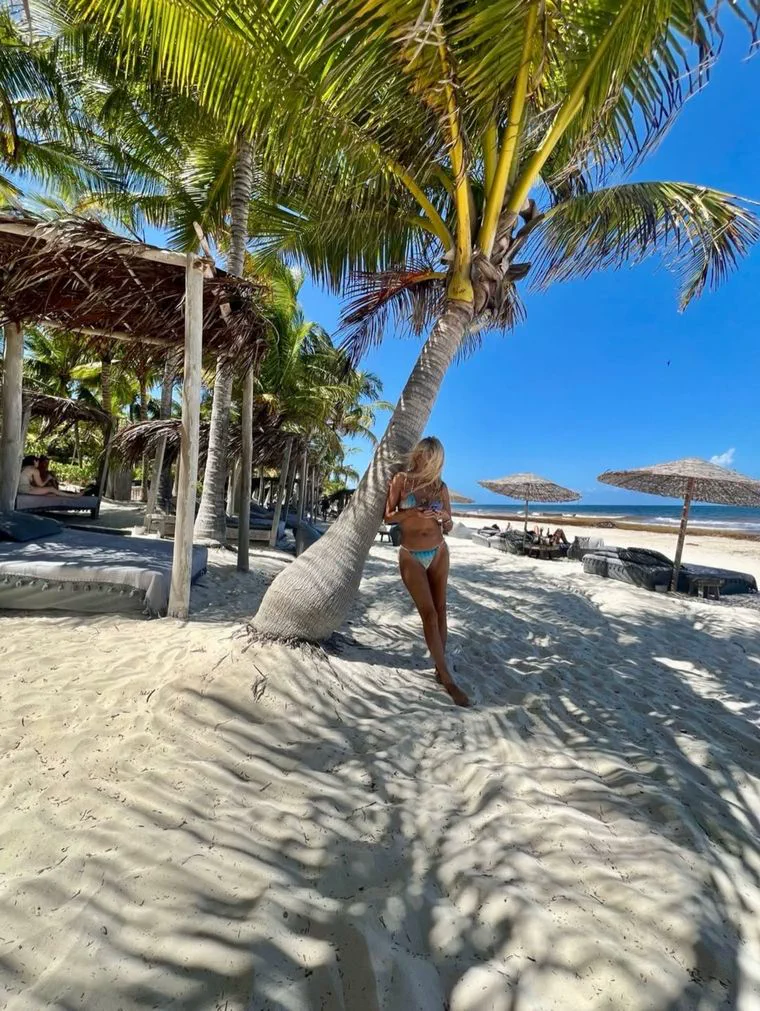 Una mujer en bikini apoyada sobre el tronco inclinado de una palmera en una playa de arena clara.