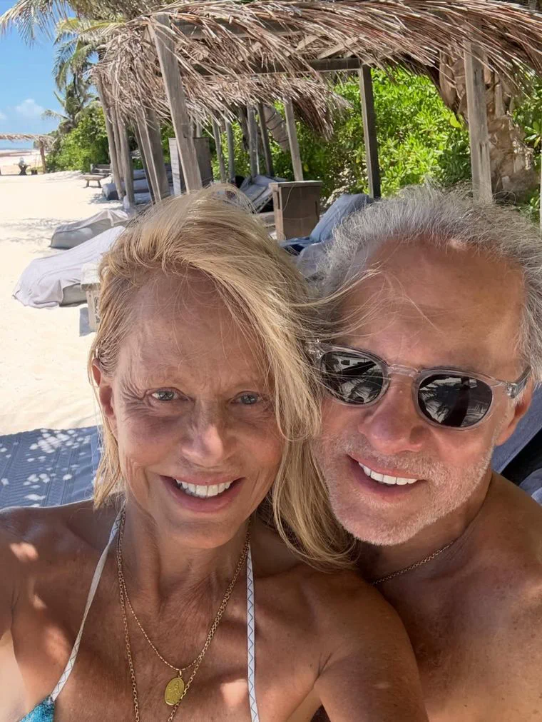 Una mujer rubia y un hombre con anteojos de sol sonríen posando para una selfi en la playa.