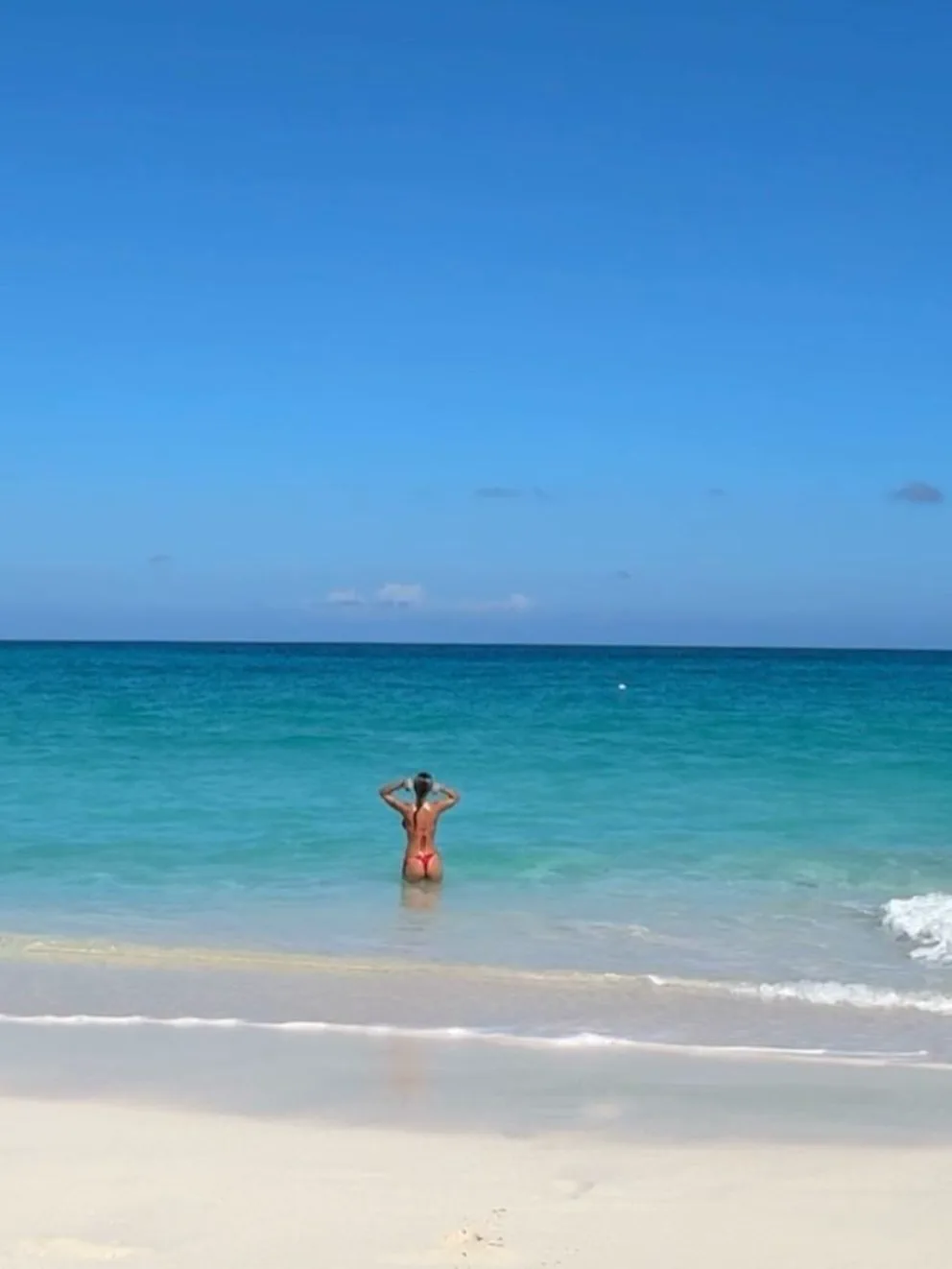 Una persona en bikini de espaldas se mete al mar con las manos en la nuca bajo un cielo despejado.