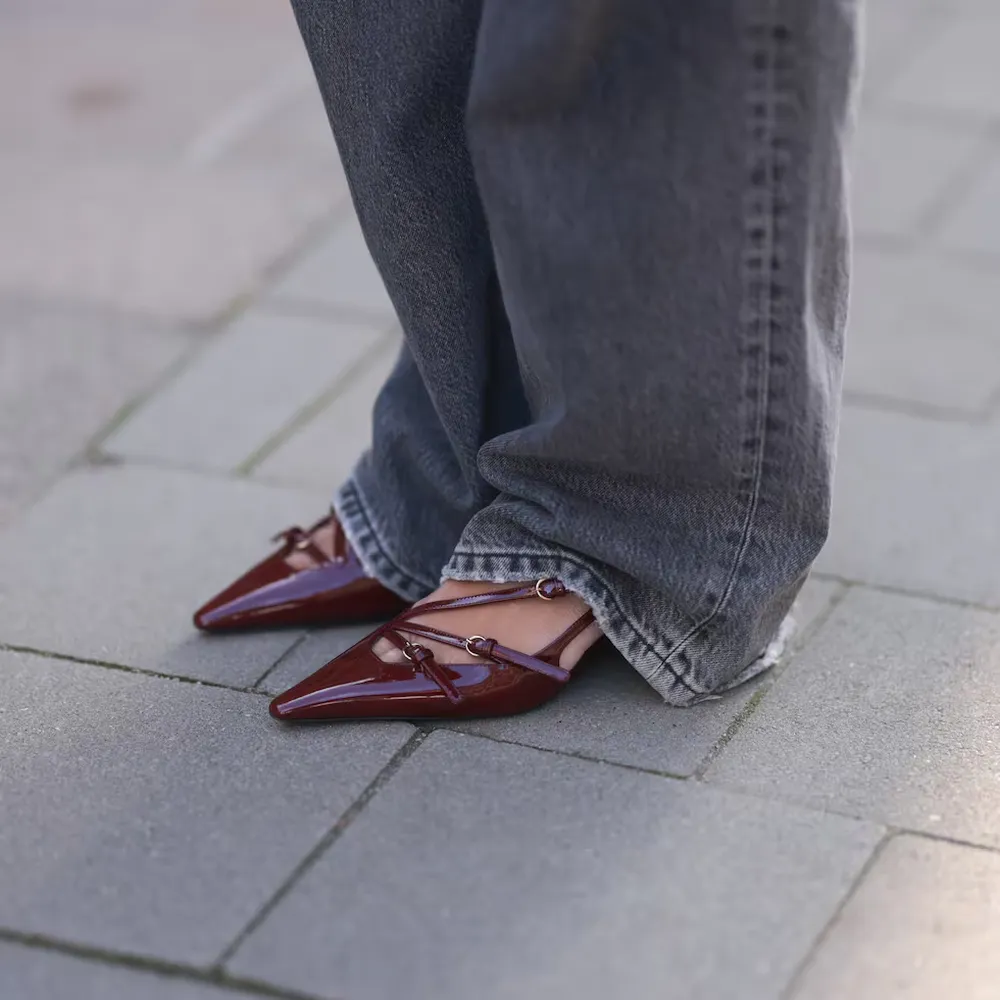 Pies de una persona con jeans anchos que lleva zapatos de charol bordó en punta con tiras finas sobre la vereda.