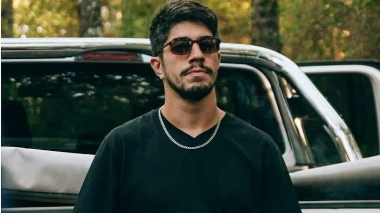 Un hombre joven con barba, anteojos de sol, remera negra y cadena plateada posa delante de una camioneta.