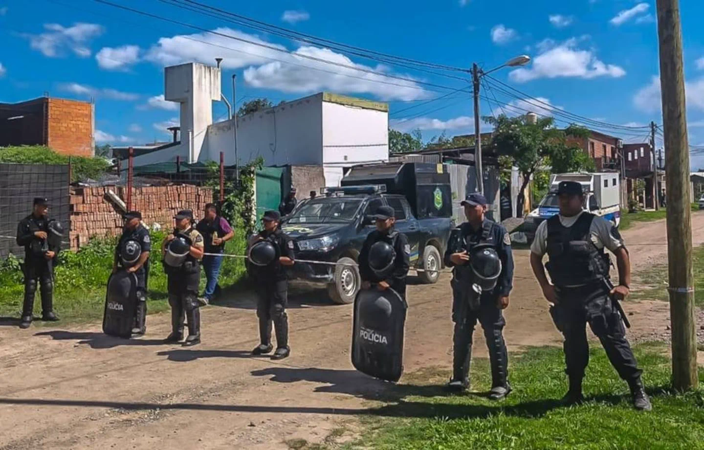 Efectivos policiales armados y con escudos forman un cordón en una calle de tierra frente a patrulleros.