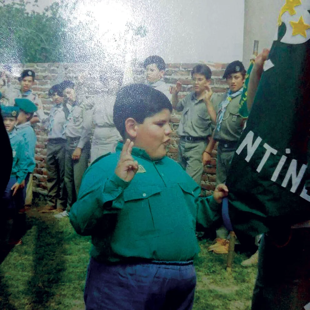 Un nene scout vestido de verde hace la seña con la mano en alto junto a una bandera y otros compañeros.