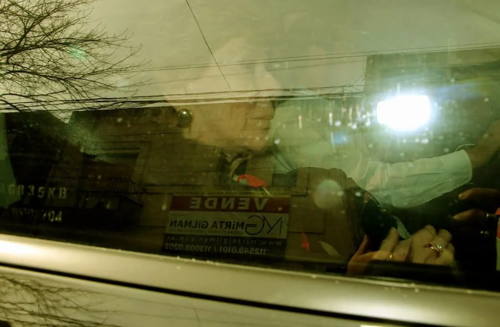 Una mujer mayor sentada dentro de un auto es vista de perfil a traves de la ventanilla con reflejos de la calle.