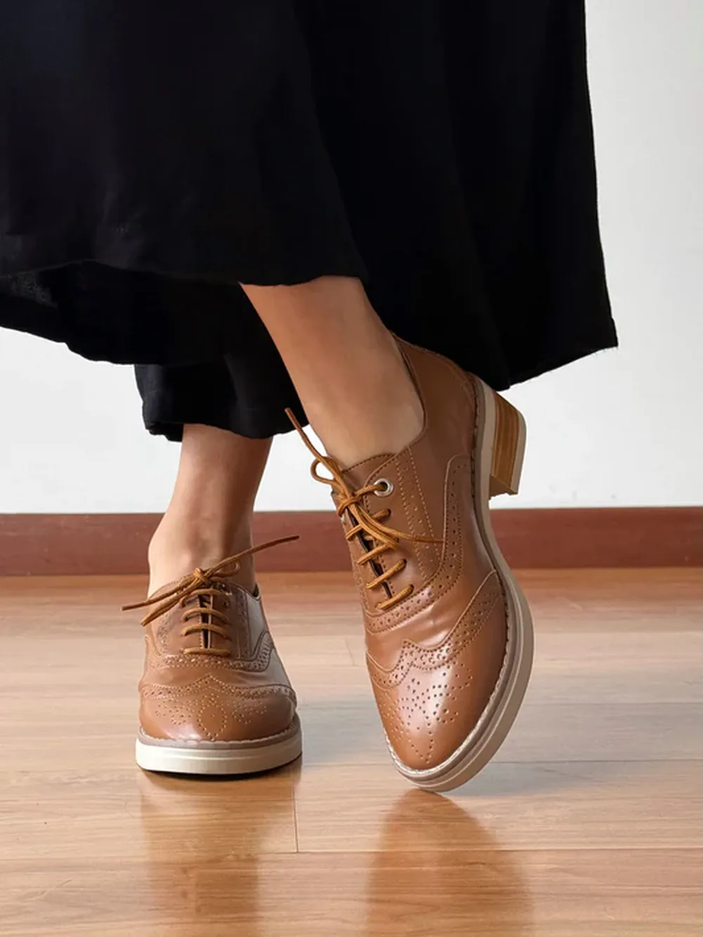 Pies de una persona sobre piso de madera usando zapatos de cuero marrones con cordones y detalles calados.