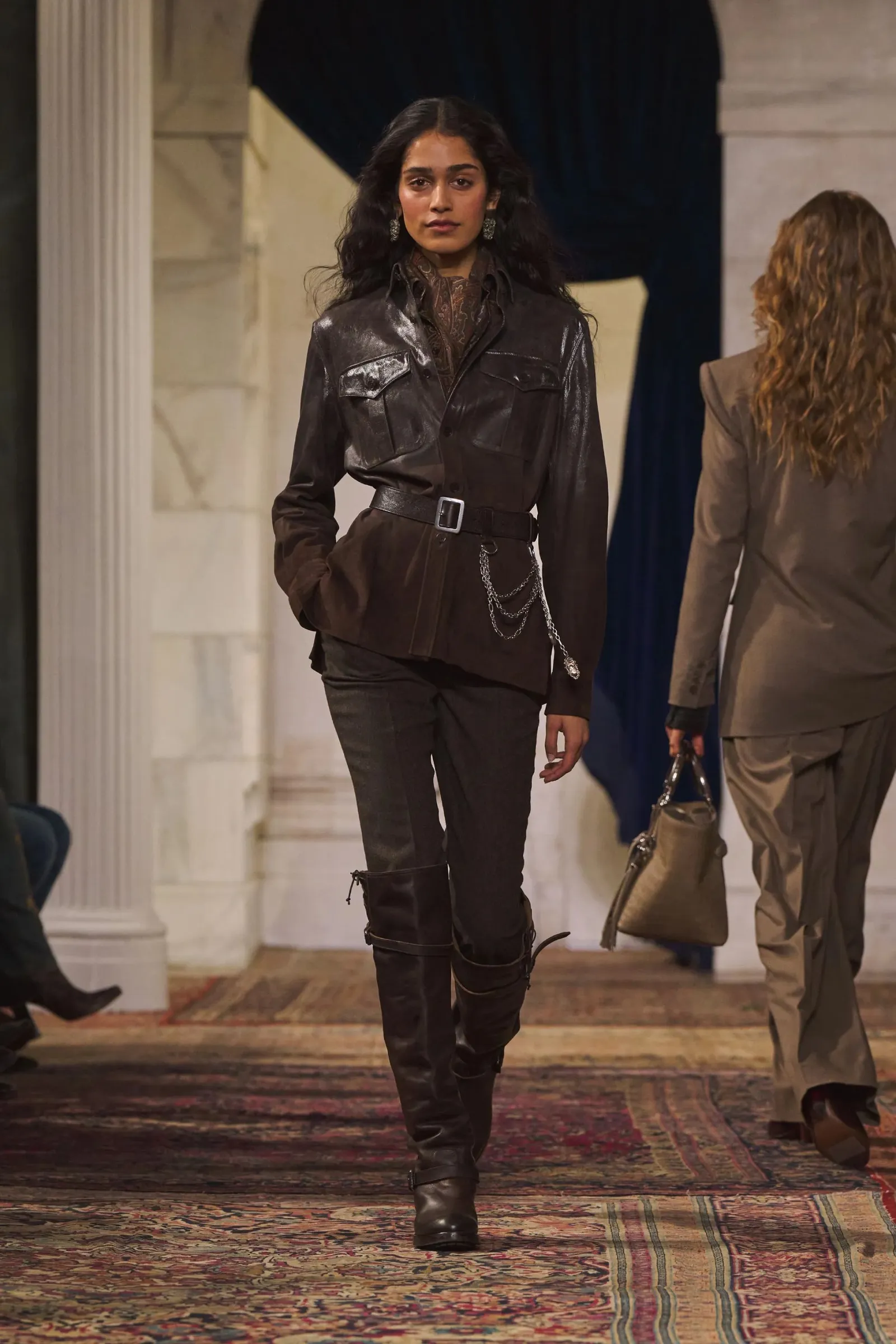 Una modelo desfila con campera de cuero marrón, cinturón ancho, pantalón y botas altas.