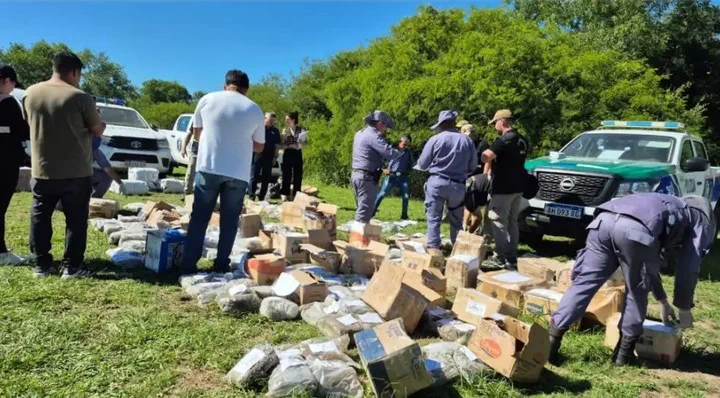 Varios efectivos de seguridad y civiles revisan y custodian decenas de paquetes apilados sobre el pasto al aire libre.