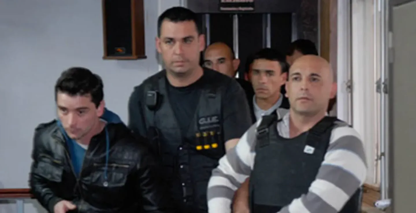 Varios hombres caminan en un interior escoltados por personal de seguridad con chalecos antibalas.