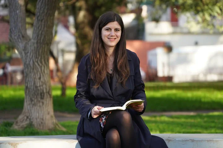 Carola della Paolera esta sentada en un banco al aire libre con un libro abierto apoyado sobre la falda.