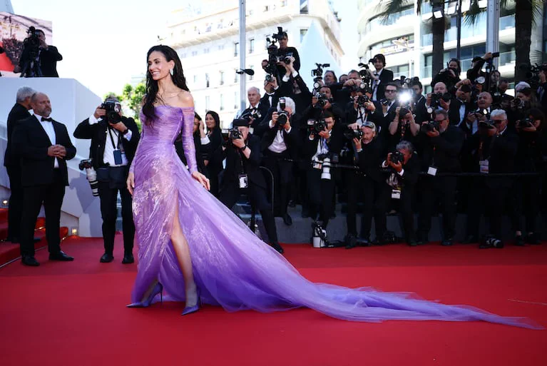 Demi Moore camina con un vestido lila de falda amplia y tajo sobre una alfombra roja rodeada de fotógrafos.