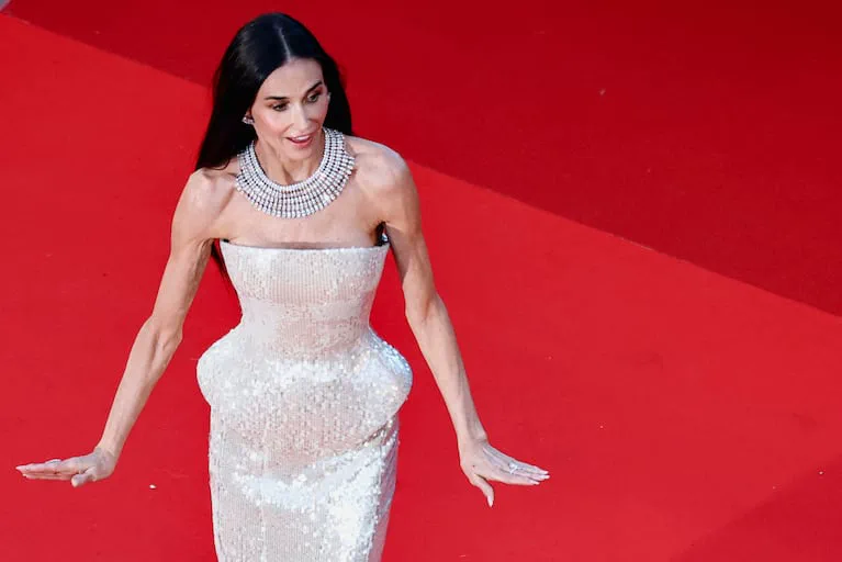 Demi Moore posa sobre una alfombra roja con un vestido blanco brillante y un collar llamativo.