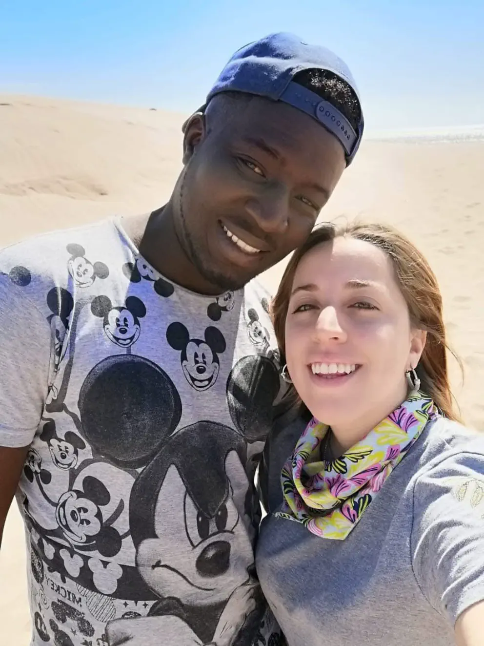 Un hombre con remera de Mickey y gorra posa sonriendo junto a una mujer en una zona de dunas de arena.