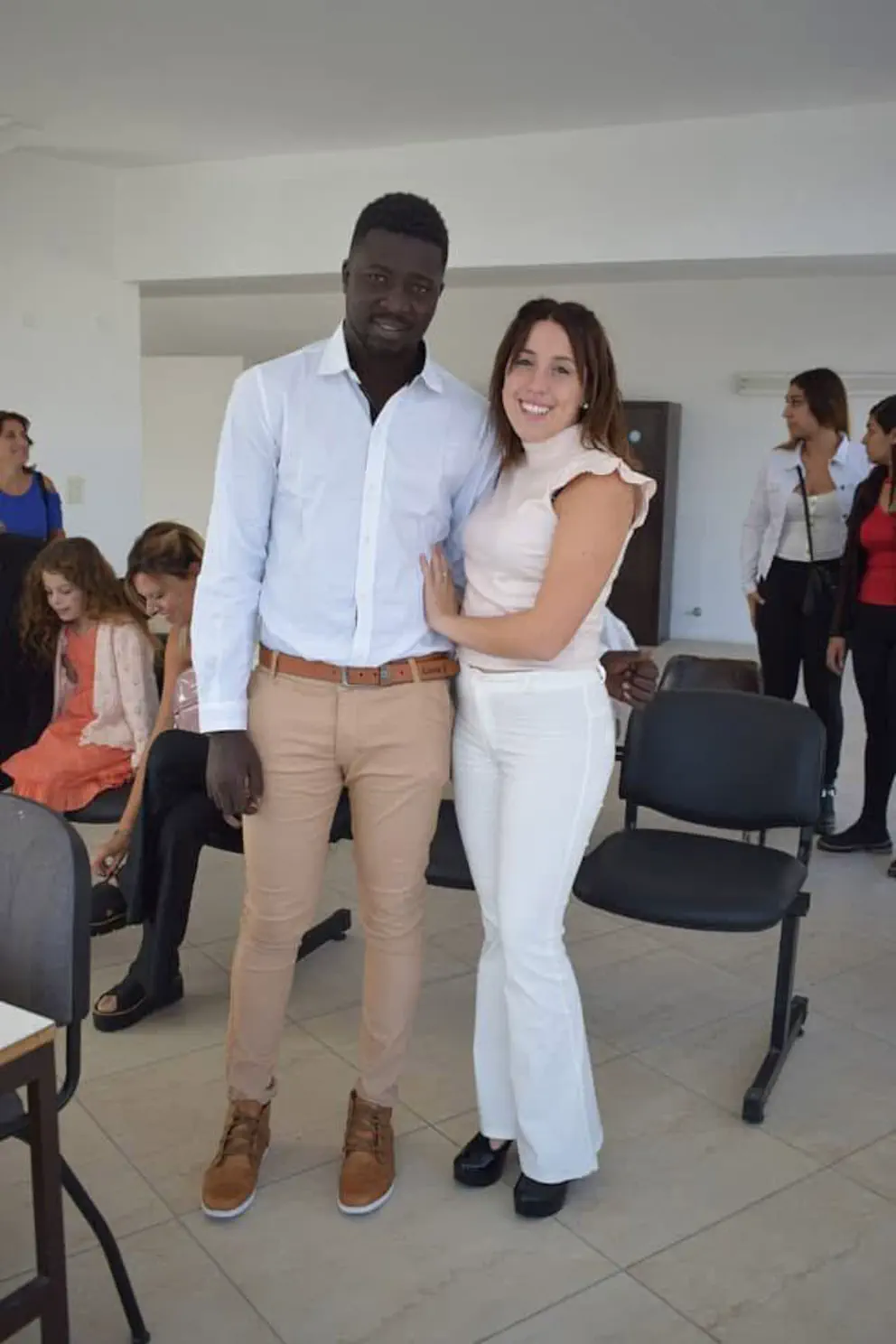 Un hombre con camisa blanca y una mujer vestida de blanco posan abrazados de pie en un salón iluminado.