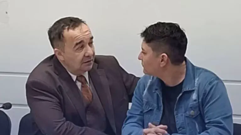 Dos hombres sentados juntos conversando de cerca, uno de traje marrón y el otro con campera de jean.
