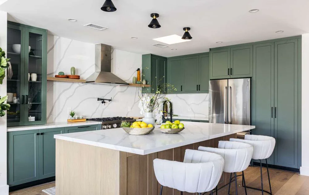 Una cocina moderna con muebles verdes, isla de mármol blanco y banquetas tapizadas.