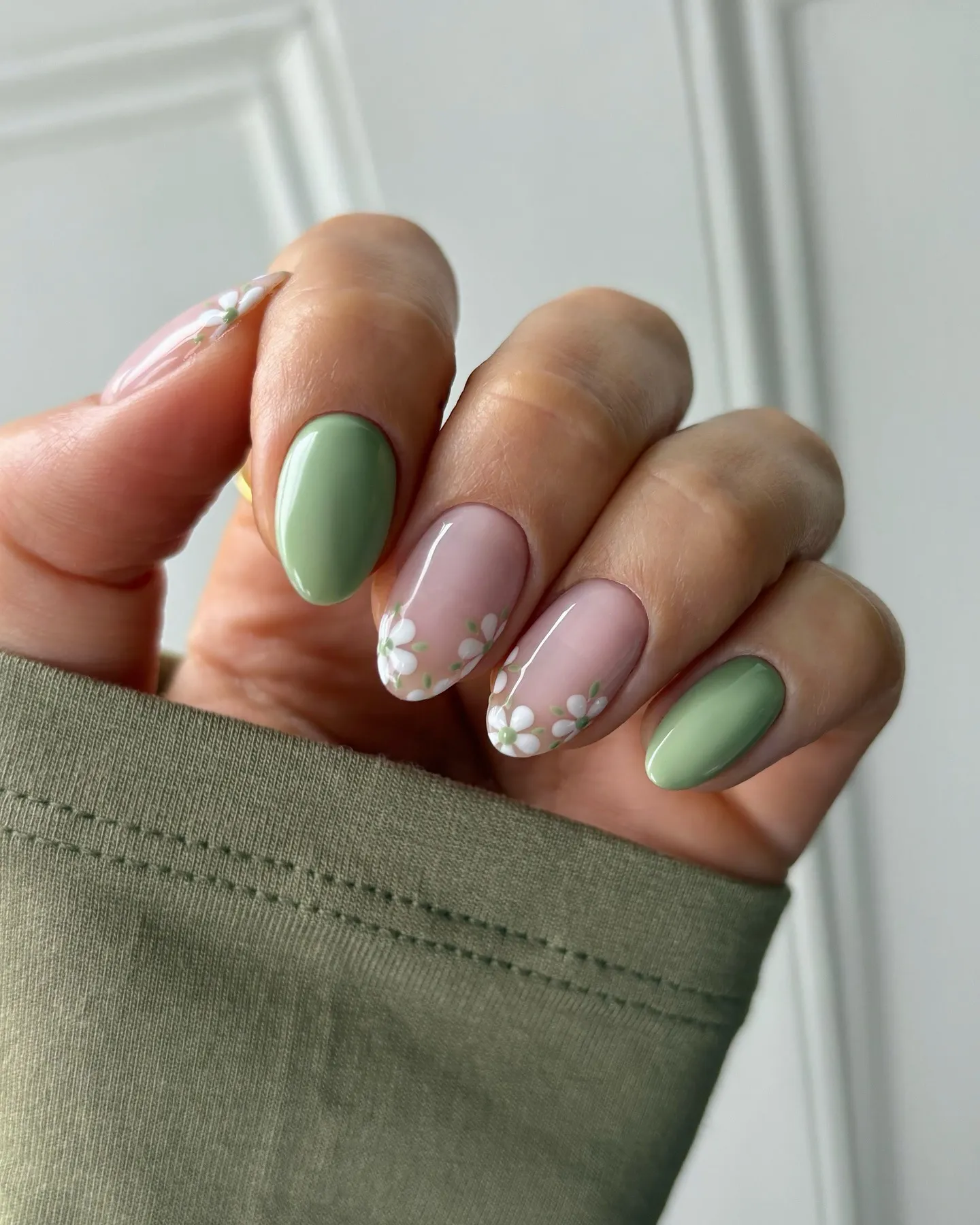 Una mano luciendo uñas pintadas de verde y tonos claros con dibujos de flores blancas.
