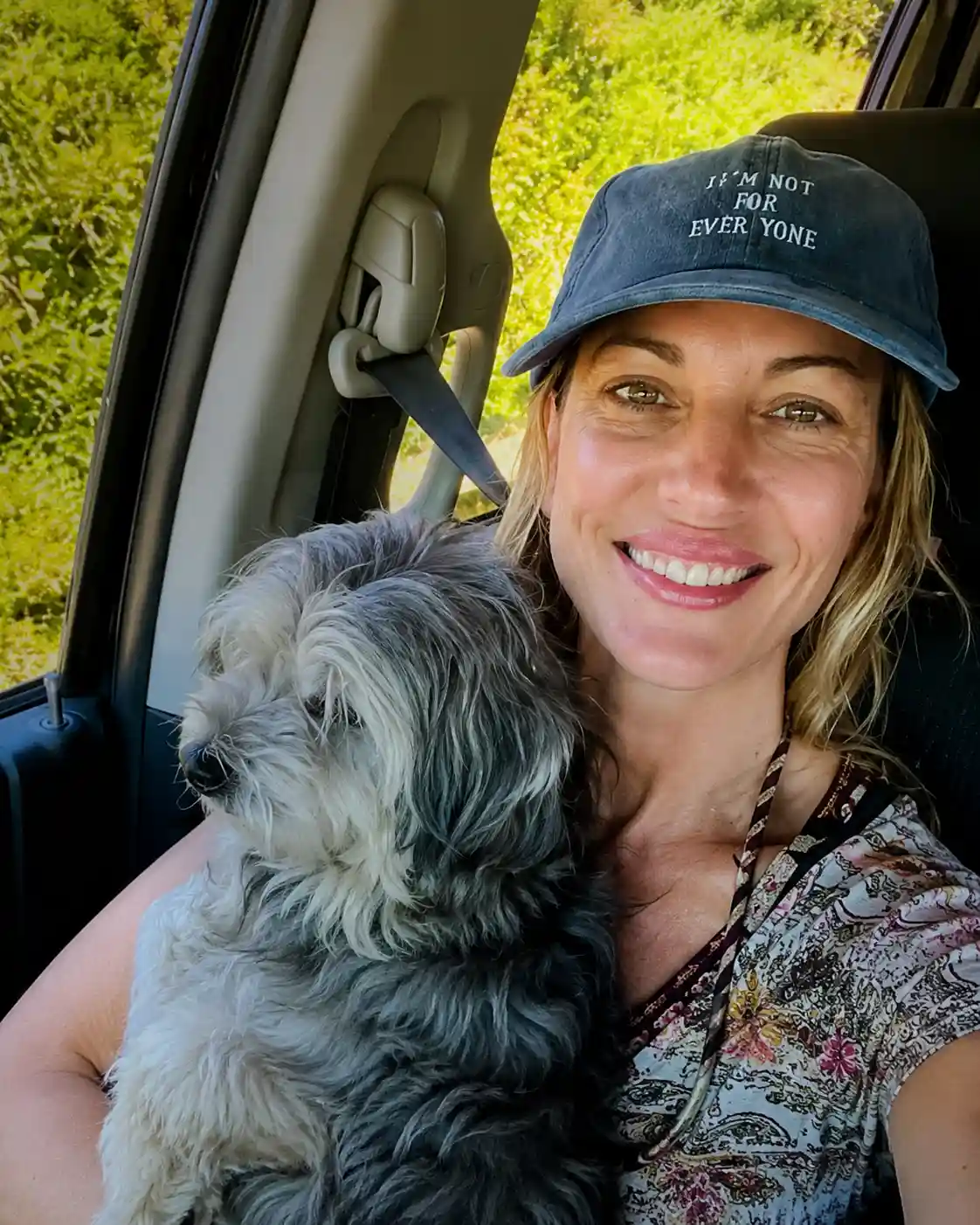 Una mujer con gorra sonríe dentro de un auto mientras sostiene a un perro gris en sus brazos.