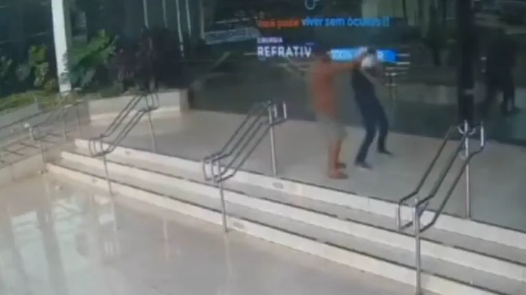 Dos personas forcejean en lo alto de una escalinata frente a la fachada vidriada de un edificio.