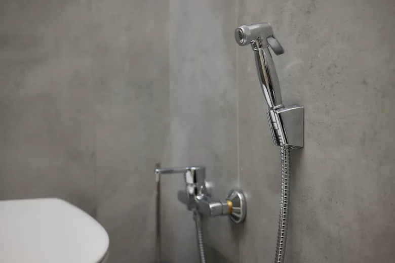 Una ducha de mano metálica con manguera cuelga de un soporte en la pared al lado de un inodoro.