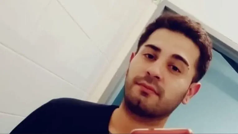 Un joven con barba corta y remera oscura mira hacia abajo mientras sostiene un celular frente al espejo.