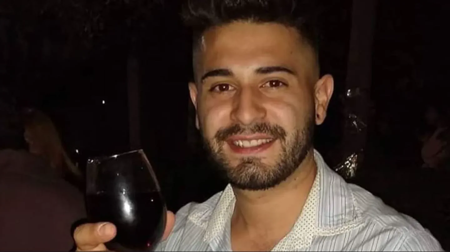 Un joven con barba y camisa a rayas sonríe mientras sostiene una copa con vino tinto en la mano.
