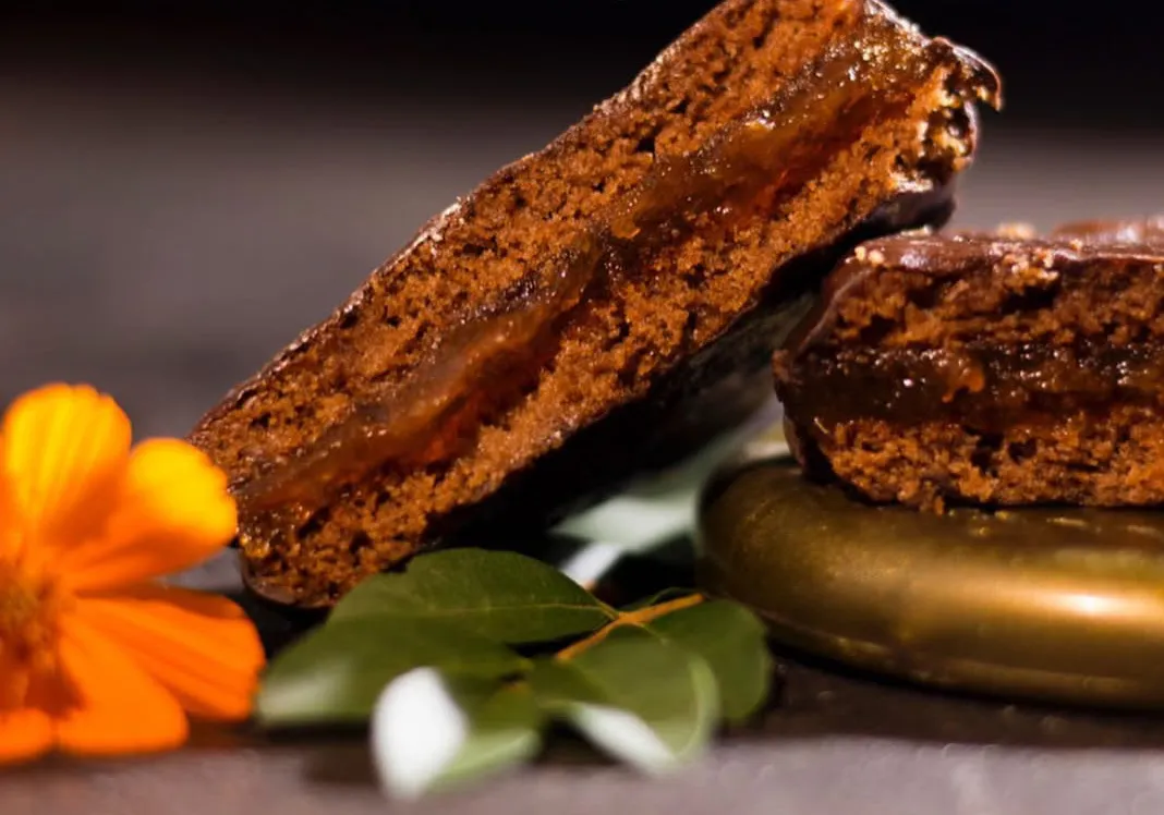 Un alfajor de chocolate relleno cortado al medio junto a unas hojas verdes y una flor naranja.