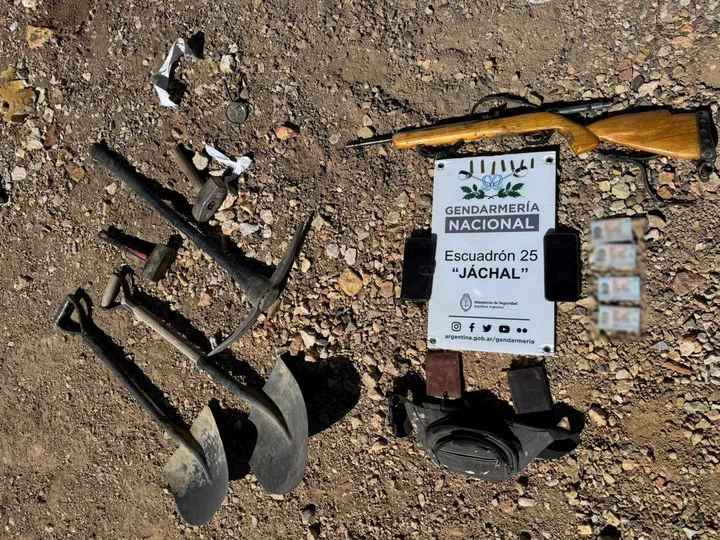 Herramientas de excavacion, un rifle, celulares, documentos y pertenencias apoyados sobre un terreno pedregoso.
