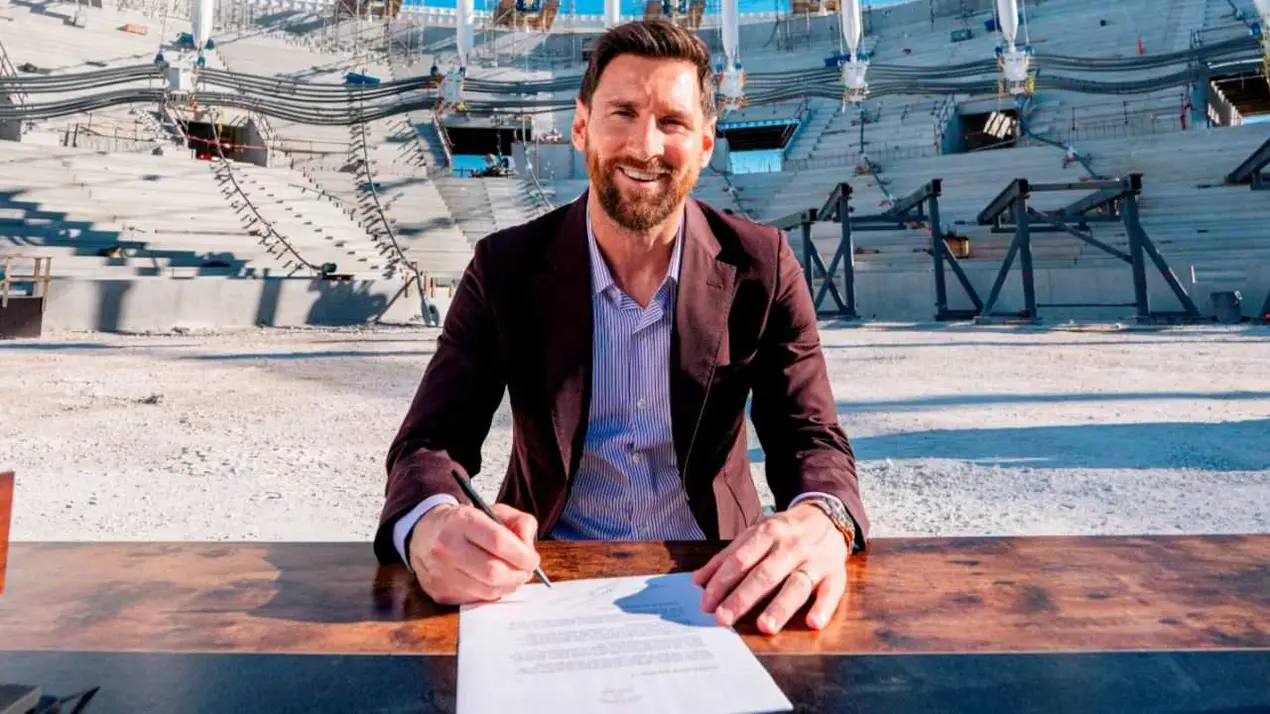 Lionel Messi sentado a una mesa firmando un documento frente a una tribuna en construccion.
