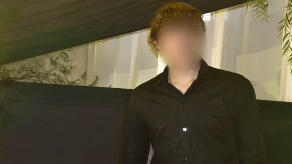 Un pibe con camisa negra posa frente a una cortina clara y una pared oscura con el rostro difuminado.