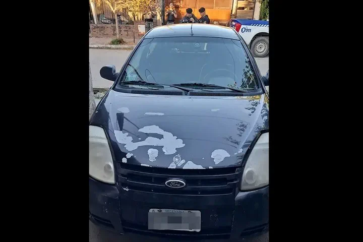 Un auto negro estacionado con la pintura del capó desgastada y policías de fondo.