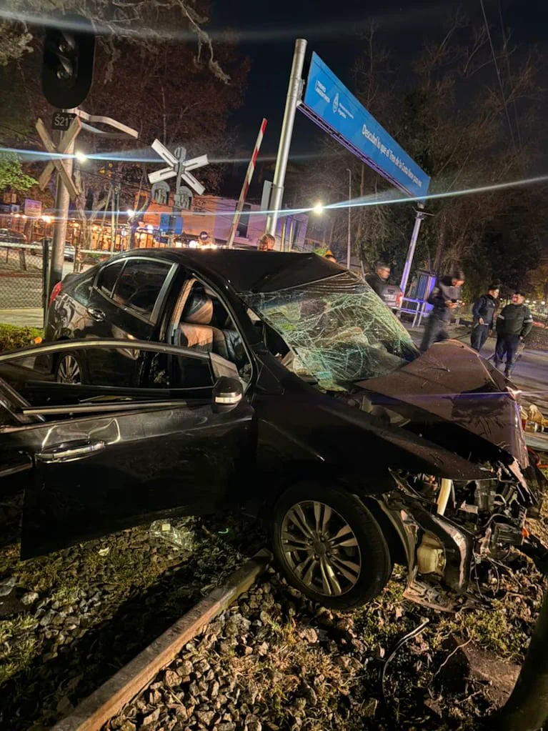 Un auto negro chocado y con el parabrisas destrozado está sobre las vías de un tren de noche.
