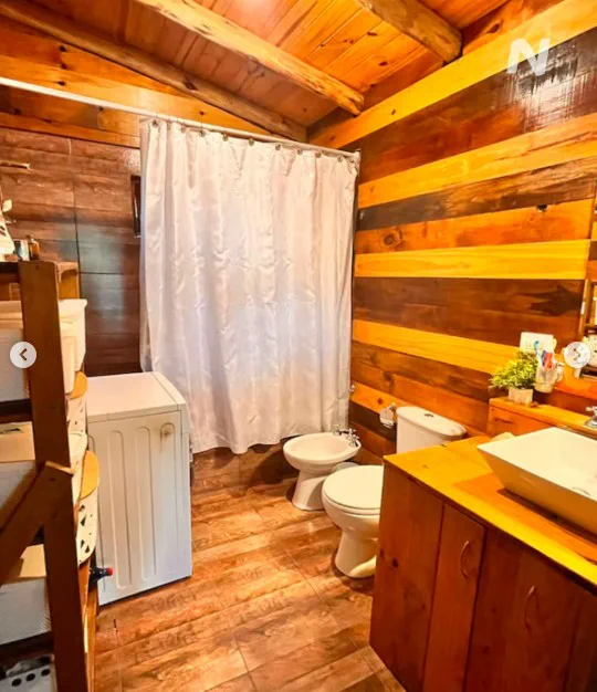 Bano revestido en madera con lavamanos, inodoro, bidet, lavarropas y cortina de ducha blanca.