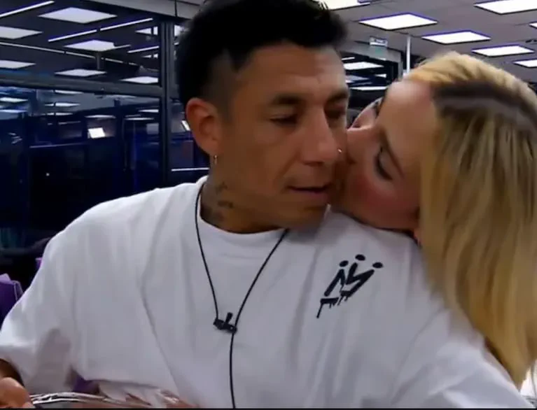 Una mujer rubia besa en la mejilla a un hombre que lleva una remera blanca con un micrófono corbatero.