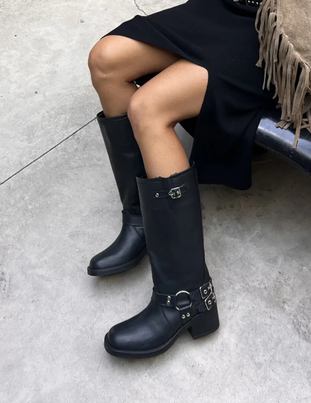 Piernas de una persona sentada que viste falda negra y botas altas de cuero negro con hebillas y argolla.