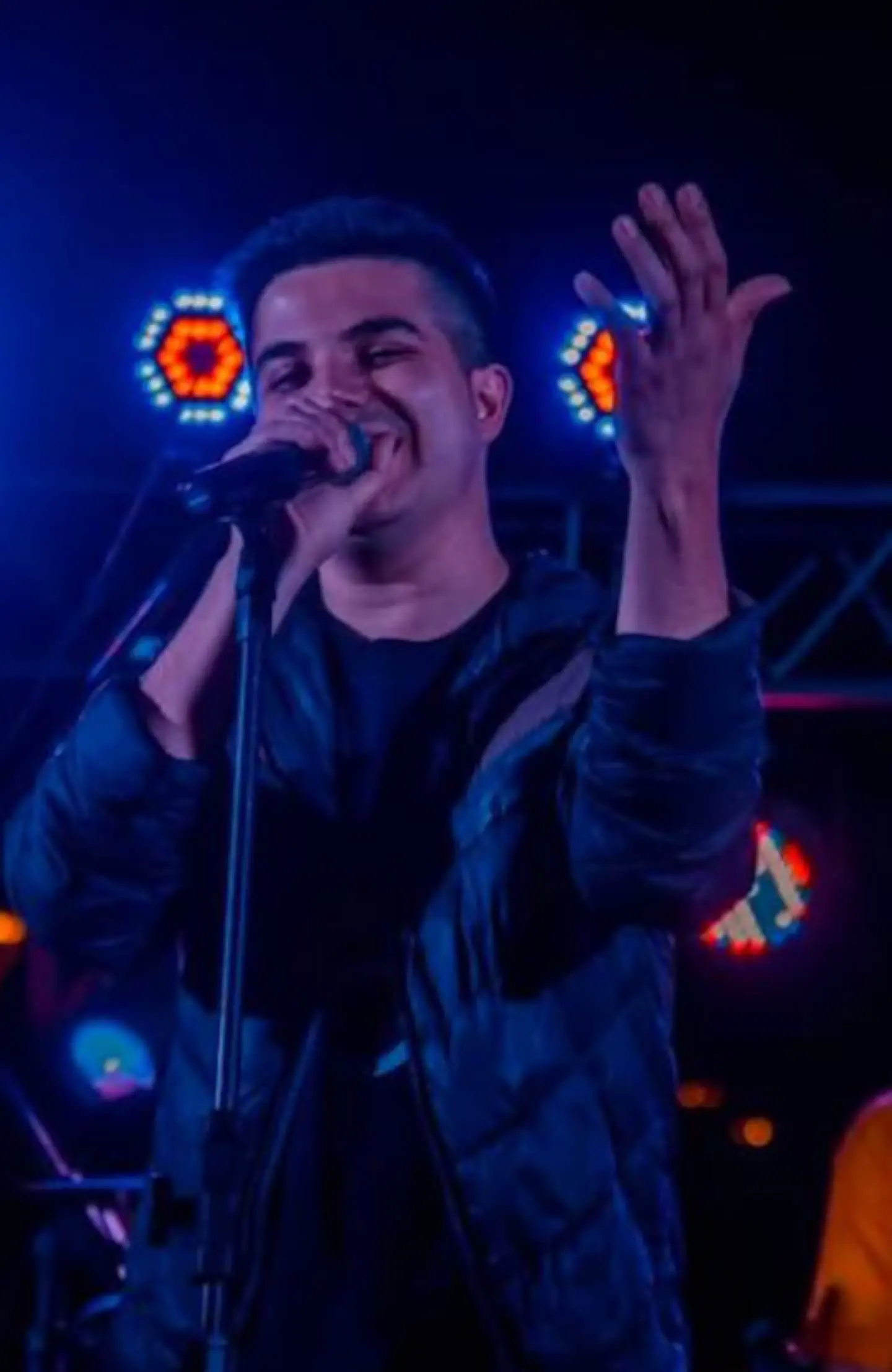 Un joven canta en un micrófono sobre un escenario iluminado con luces azules.