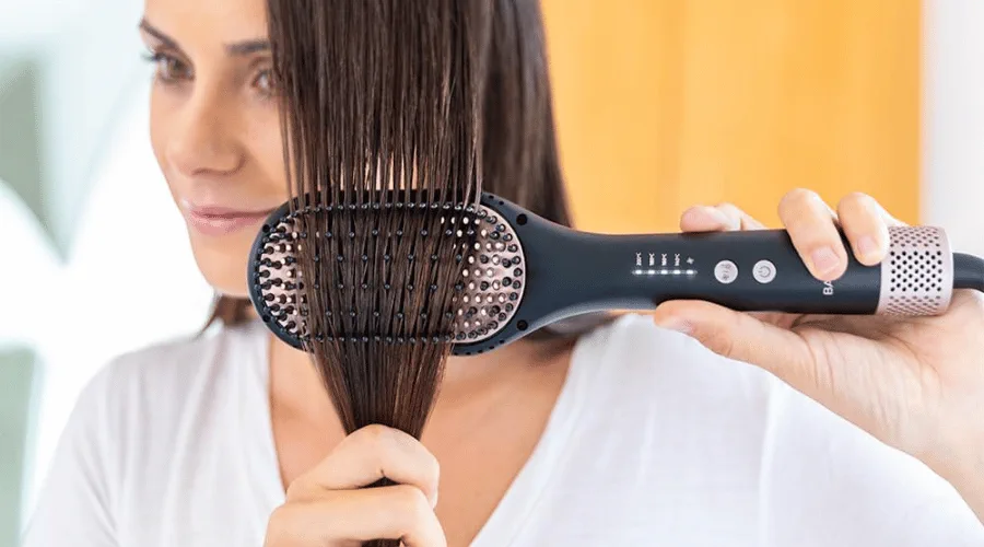 Una mujer se pasa un cepillo alisador electrico por un mechon de pelo castaño humedo.