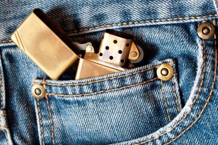Un encendedor metalico dorado abierto esta asomando en el bolsillo pequeño de un jean azul.